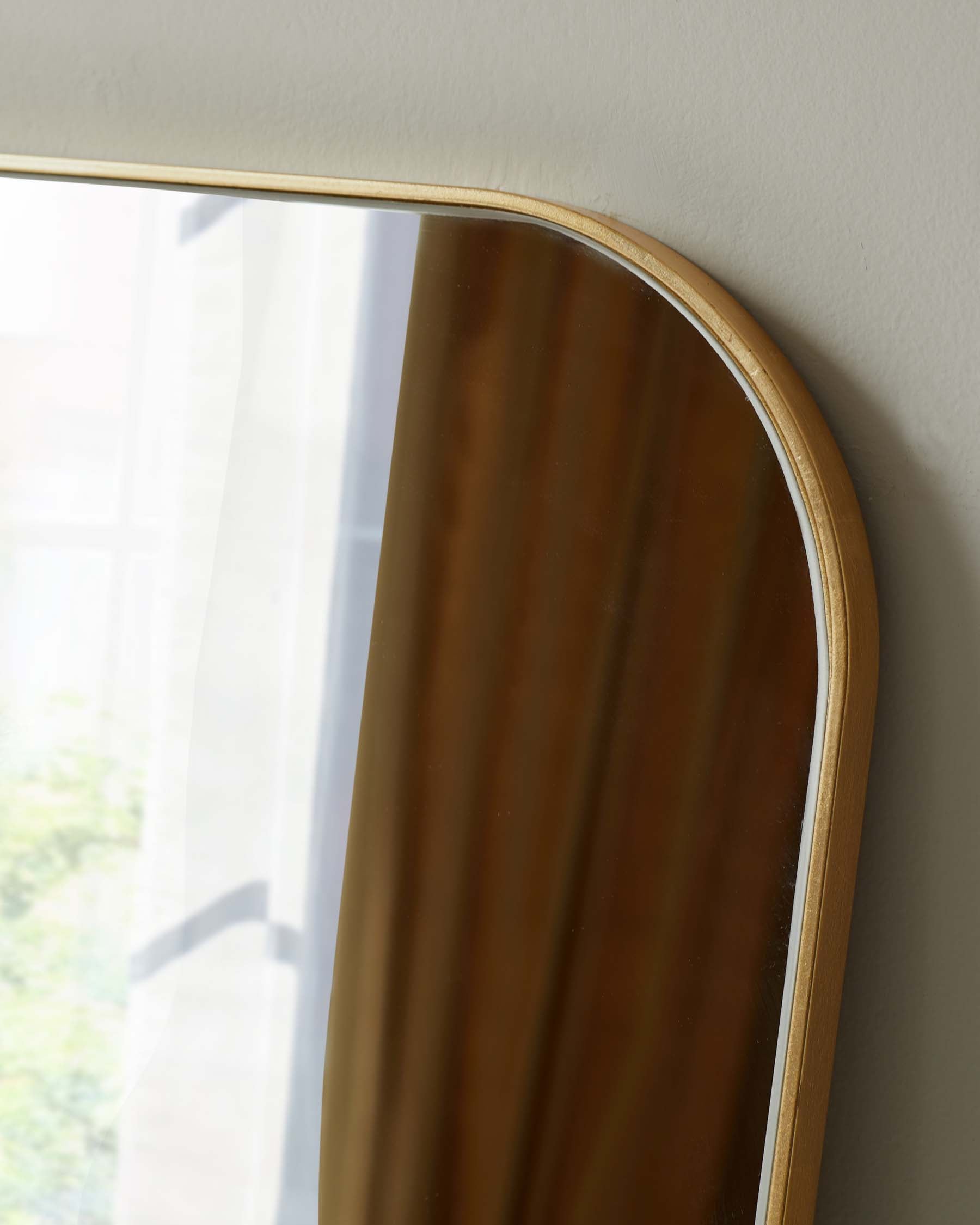 Part of a modern, rounded-edge mirror with a minimalist wooden frame reflecting a blurred view of a window and foliage.