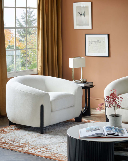 Two cozy white armchairs with black legs, a round coffee table with a sleek design, and a stylish lamp beside a window.