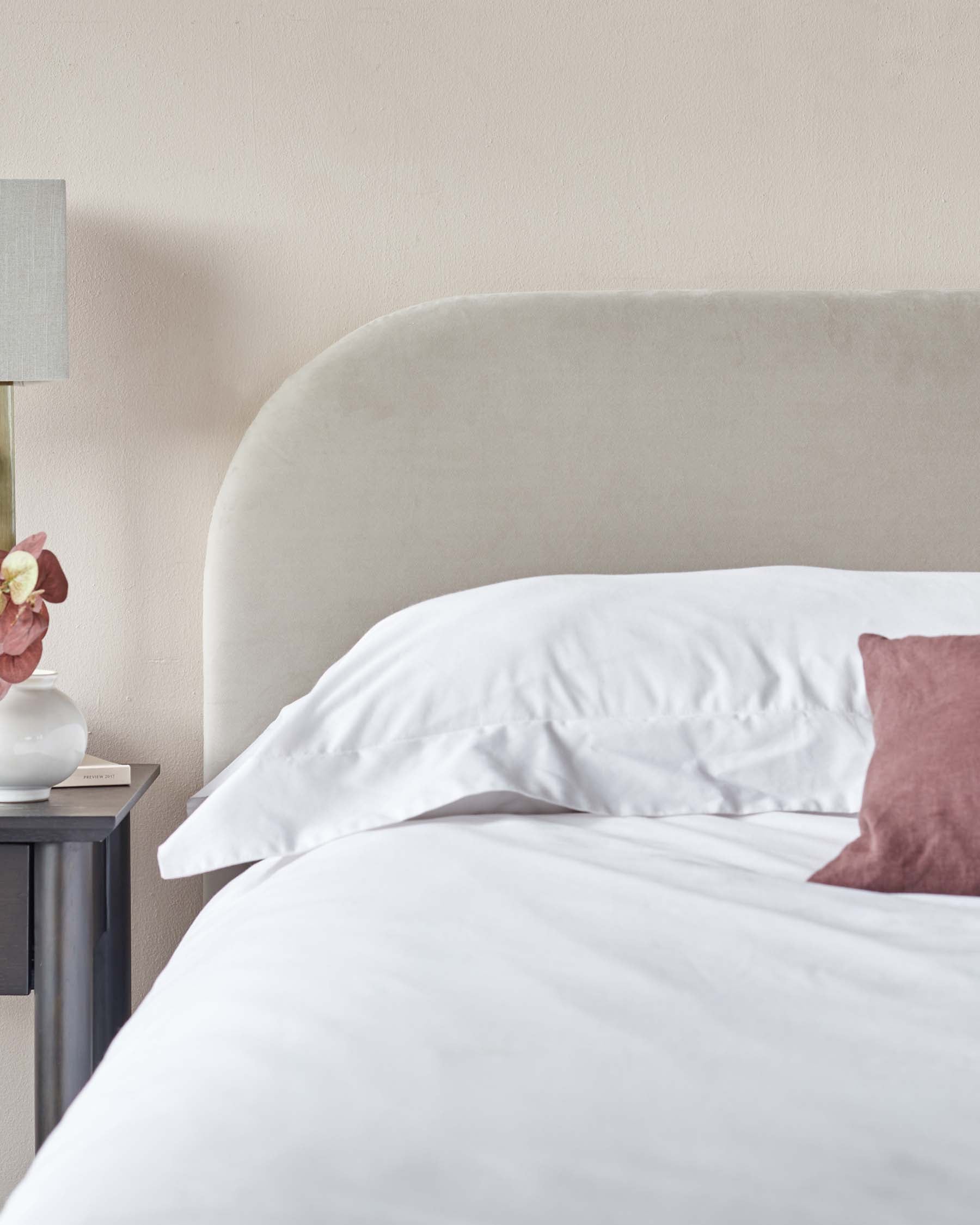 Plush beige upholstered headboard with a gentle curve on the top, crisp white bed linens, and a grey bedside table with a simple, modern design featuring a table lamp with a textured grey lampshade.