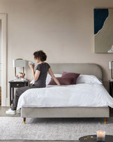 Modern bedroom featuring a minimalist upholstered bed with a curved headboard and tapered wooden legs, flanked by two black bedside tables with a classic design and brass knobs. A textured light-coloured area rug lies beneath the bed, adding warmth to the space.