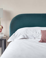 Elegant teal upholstered headboard with a smooth curved top, paired with a minimalist white bedside table featuring a single drawer, set against a soft beige wall. White bedding and a pink accent pillow complete the serene bedroom scene.