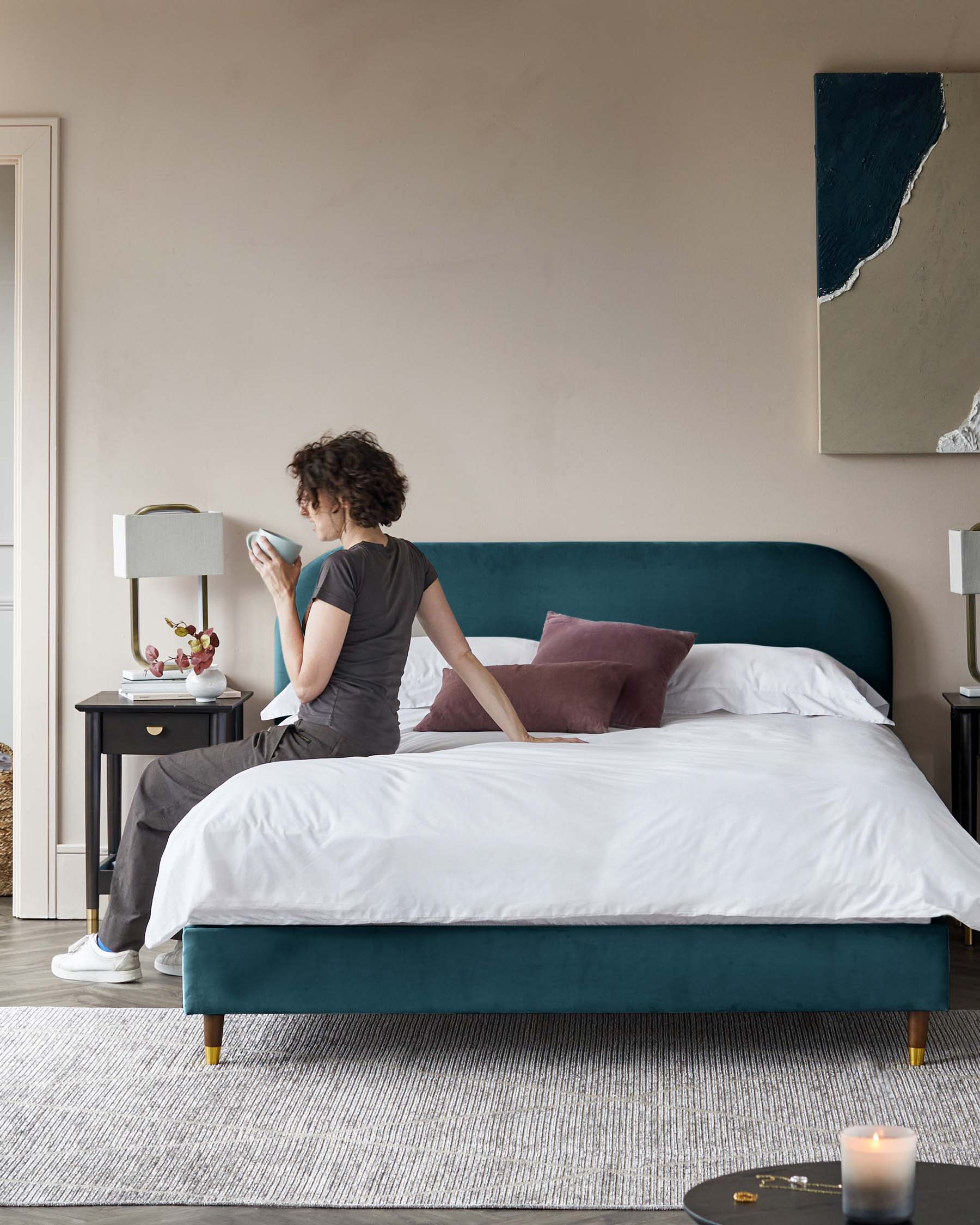 Elegant bedroom furniture featuring a plush teal upholstered bed with a curved headboard and gold-capped legs, flanked by two classic black bedside tables with gold handles and a matching round black coffee table with a gold rim and legs. The setting is complemented by a textured off-white area rug under the bed.