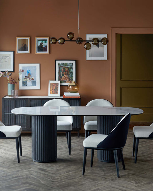 Elegant oval dining table with a ribbed column base, complemented by modern, upholstered chairs in soft tones and a stylish pendant light.