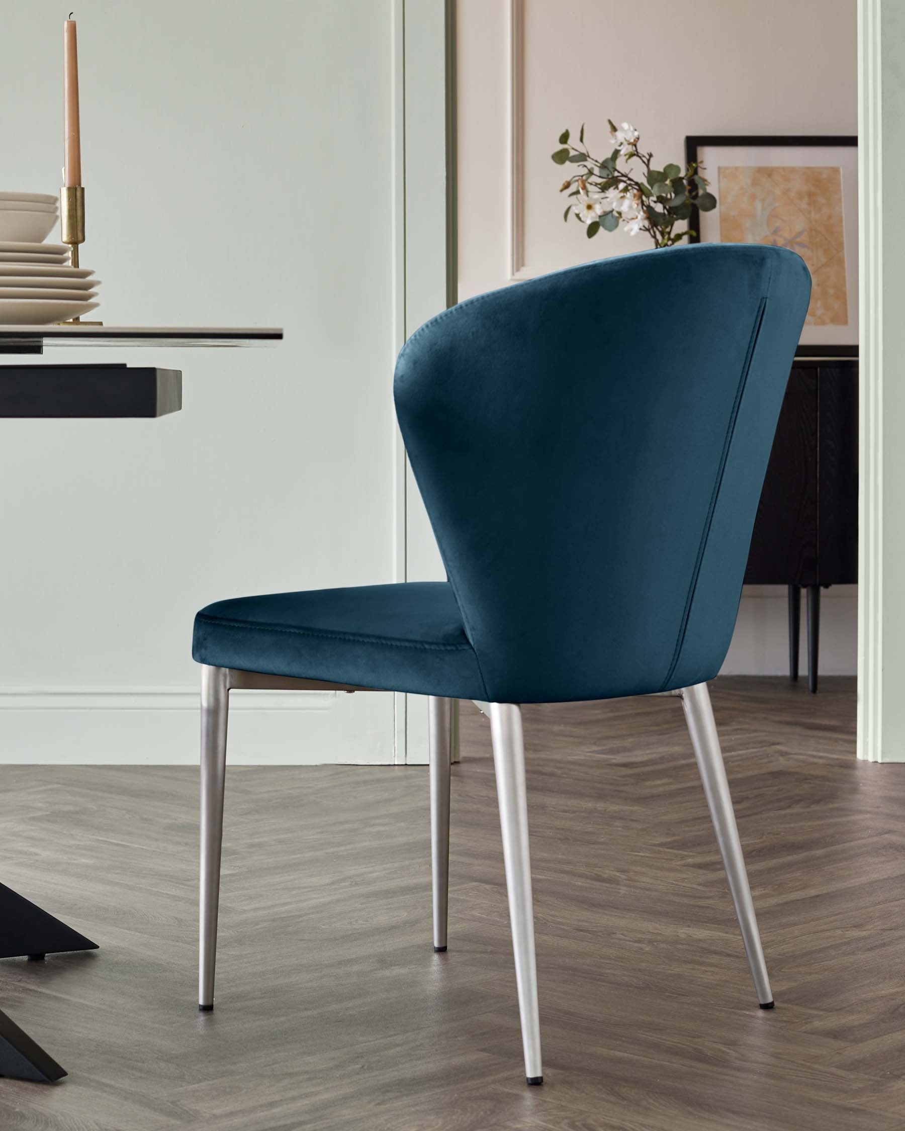 Elegant modern dining chair with plush teal blue velvet upholstery and sleek, tapered silver metal legs. The chair has a curved backrest for added comfort and a stylish silhouette that brings a touch of sophistication to any dining room setting.