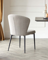 Modern minimalist accent chair with a curved backrest, upholstered in a light grey textured fabric, complemented by sleek, angled black metal legs. The chair is showcased in a bright, elegant room with a wooden floor, placed adjacent to a wall-mounted dark wooden shelf with decorative objects.