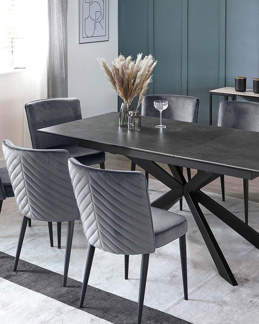 Modern dining set featuring a dark table with an angular base and elegant gray velvet chairs, complemented by a decorative vase.