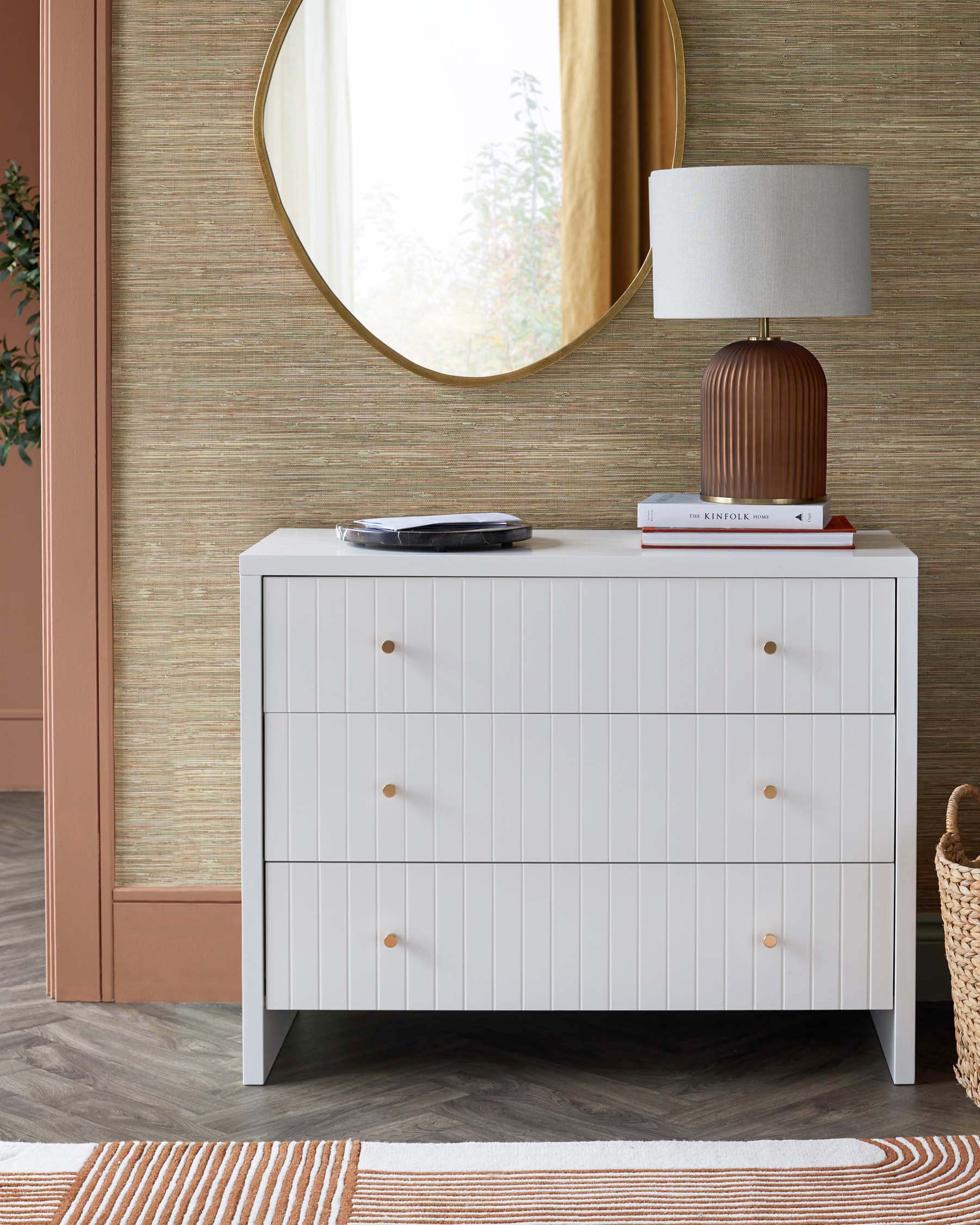 A modern white dresser with vertical grooves, three drawers, golden knobs, a stylish lamp, and a round mirror.