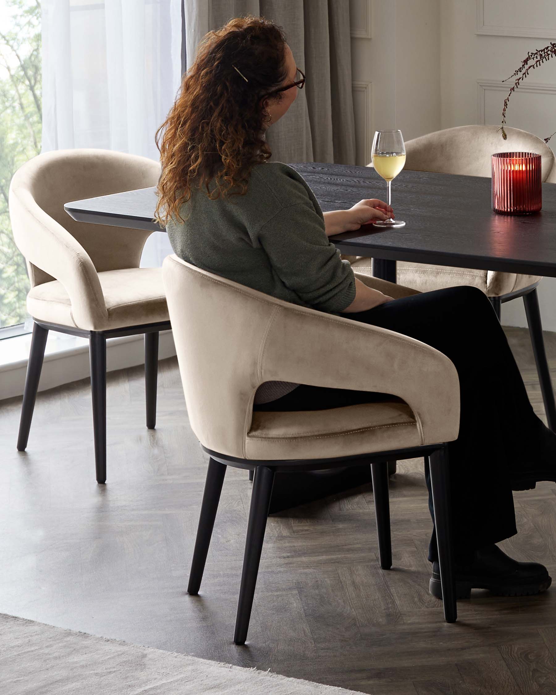 Elegant contemporary dining room furniture, featuring a round, black, wooden table with a textured top and inclined legs. Accompanied by two beige upholstered chairs with a smooth, curved design and black wooden legs, providing a sophisticated contrast.