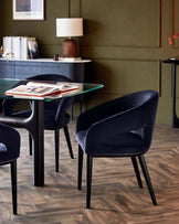 A sleek glass-top table paired with stylish navy blue upholstered chairs, set against a rich green wall and wooden flooring.