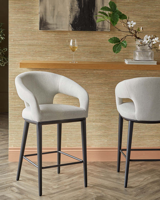 Two modern bar stools with curved backs, upholstered in light fabric, set against a textured wall and wooden shelf.