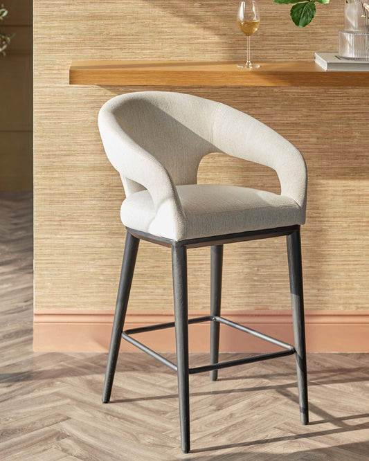 Stylish upholstered bar stool with a curved back, soft light fabric, and sturdy black legs, ideal for modern interiors.