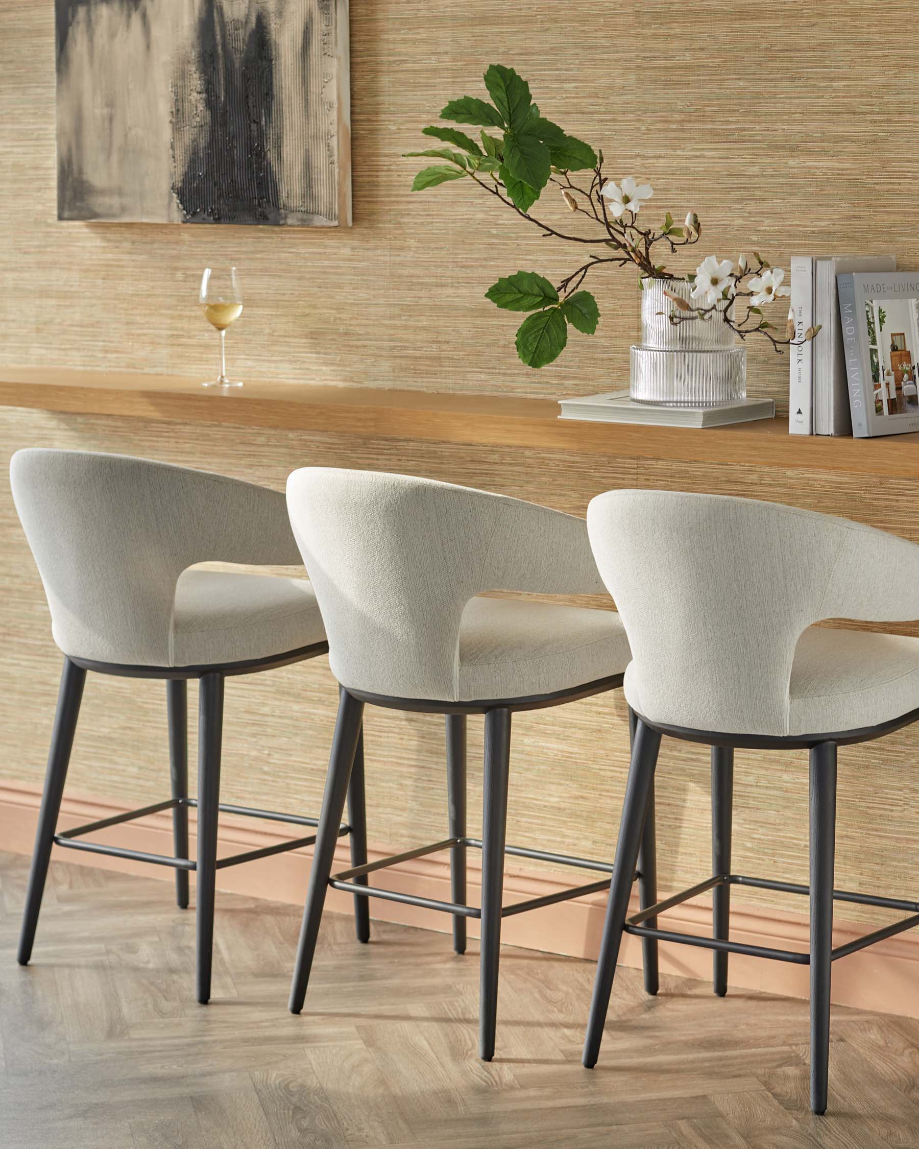 Three modern bar stools with soft, curved backs and metal legs, set against a textured wall and a wooden counter.