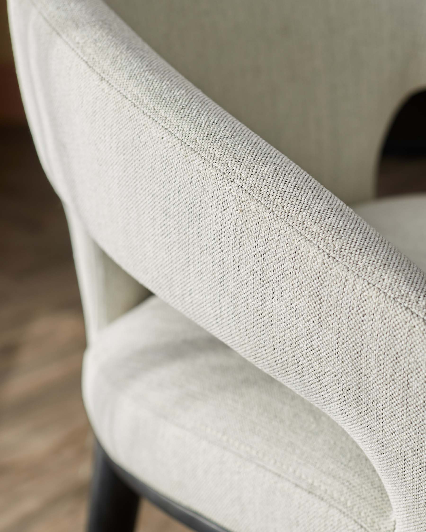 A close-up view of a stylish armchair with soft, textured light fabric and curved backrest, resting on a dark wooden base.