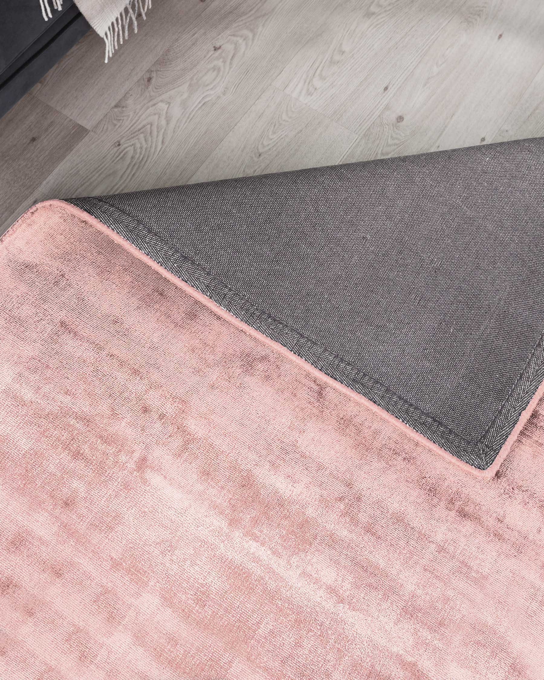 Soft-textured, blush pink area rug with a grey border, placed on a light wooden floor, partially displaying a corner of a grey upholstered ottoman with a fringed off-white throw blanket on top.