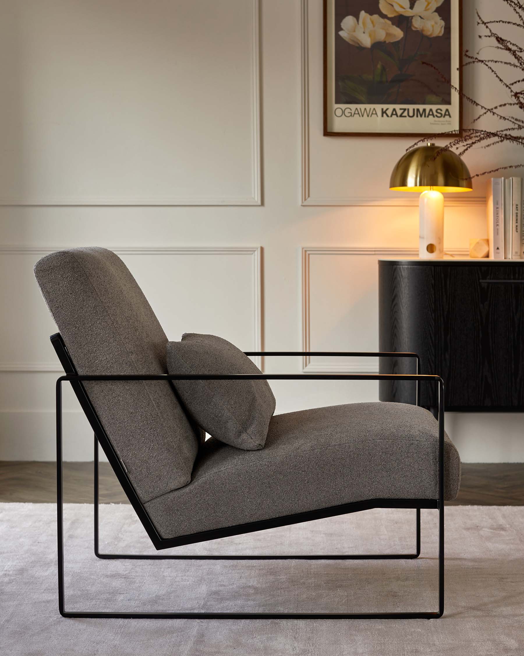 Modern grey fabric lounge chair with minimalist black metal frame on a neutral toned rug, positioned next to a sleek black sideboard with a brass coloured table lamp and framed artwork on the wall above.