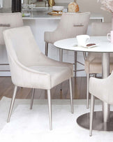 Elegant beige upholstered chairs with silver metal legs around a sleek, round white table, set in a modern dining area.