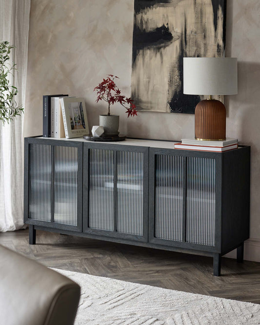 Elegant dark sideboard with ribbed glass doors, topped with a stylish lamp, decorative plant, and stacked books, in a modern setting.