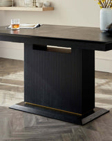 Modern dark wood dining table with a thick rectangular top and an architecturally inspired base featuring vertical fluting. The base is underscored by a gold-toned metal accent and rests on a flat, wide platform. The table surface is adorned with a glass and an open magazine. The setting is complemented by a light neutral-toned wall and light wooden flooring, suggesting a contemporary and elegant interior.