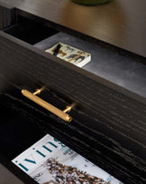 Close-up of a contemporary black wooden console table with a textured finish, featuring an open drawer with a brass handle revealing a book and another compartment with magazines.