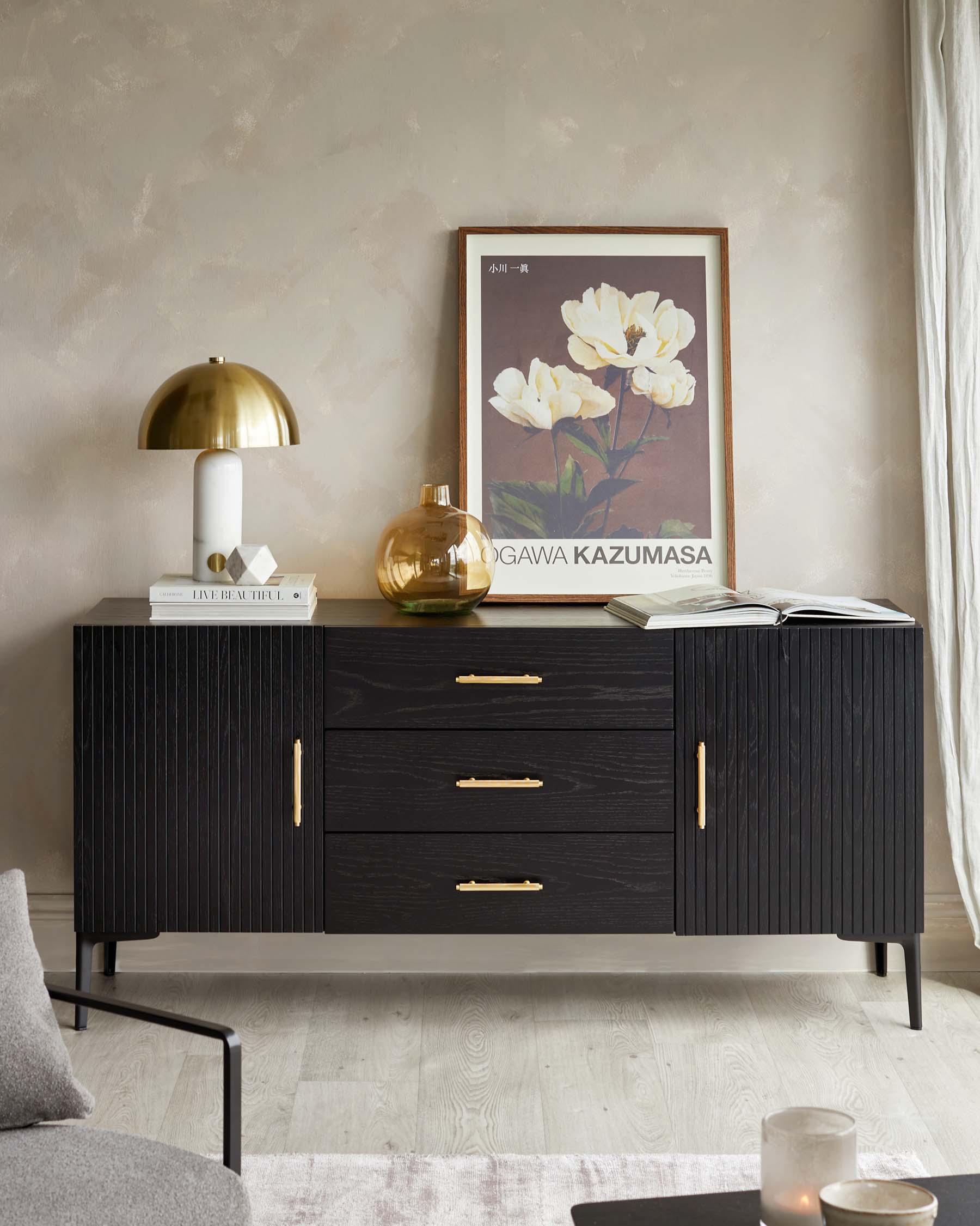 A modern, black textured wood sideboard with brass handles, featuring three drawers and a two-door cabinet, supported by slender, tapered legs. The top displays decorative items including a gold-tone lamp, vase, books, and framed artwork.