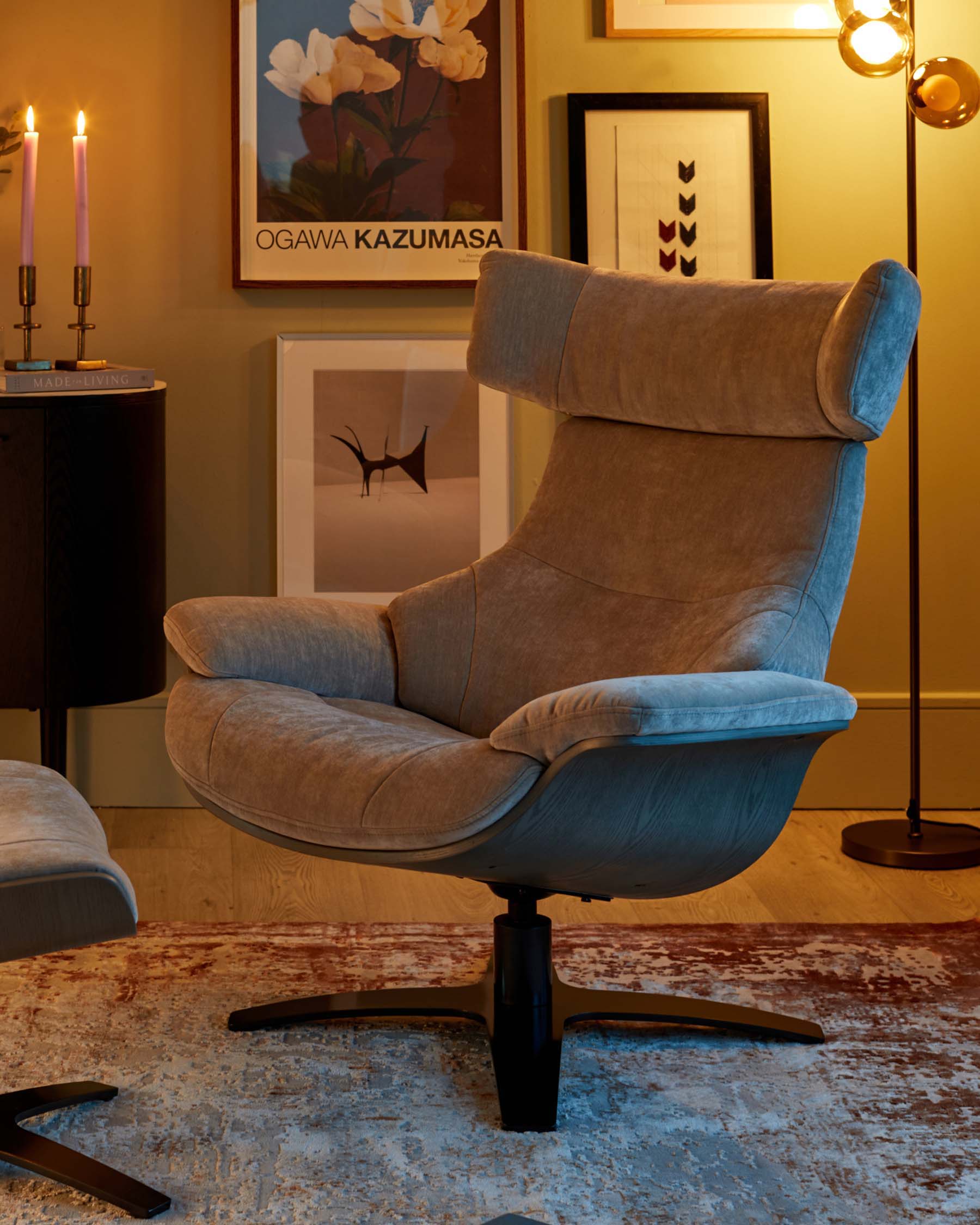 Modern recliner chair in plush grey upholstery with a contoured backrest, integrated headrest, and a padded, wrap-around seat design. The chair features a black swivel base with slender, curved footrests. A coordinating grey ottoman with a similar base is partially visible, suggesting a matching set. The scene is warmly lit to emphasize the furniture's inviting texture and contours.