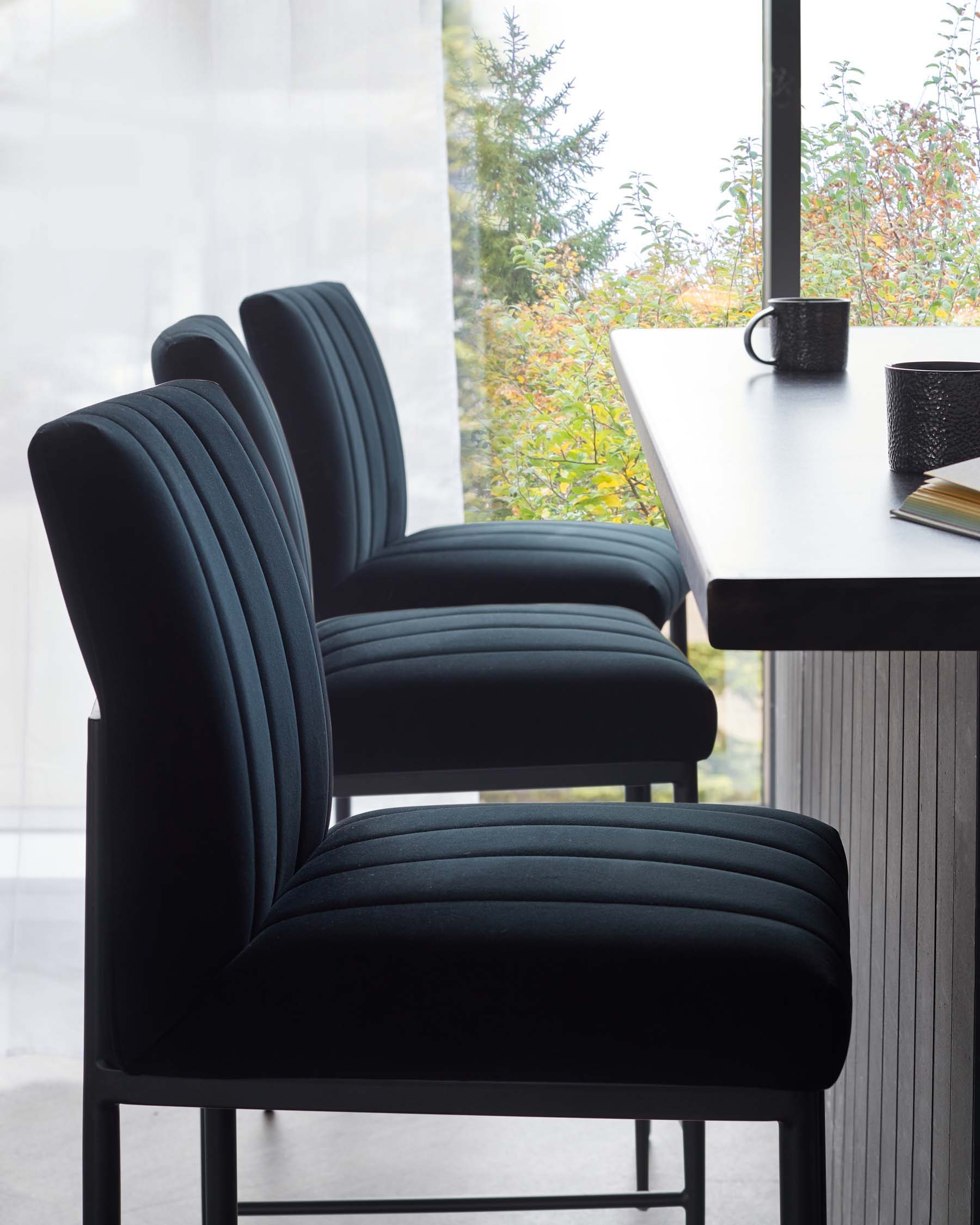 Elegant black upholstered dining chairs with vertical channel tufting and a modern curved back design, seated around a sleek white dining table, with a minimalist black mug and notebook on top.