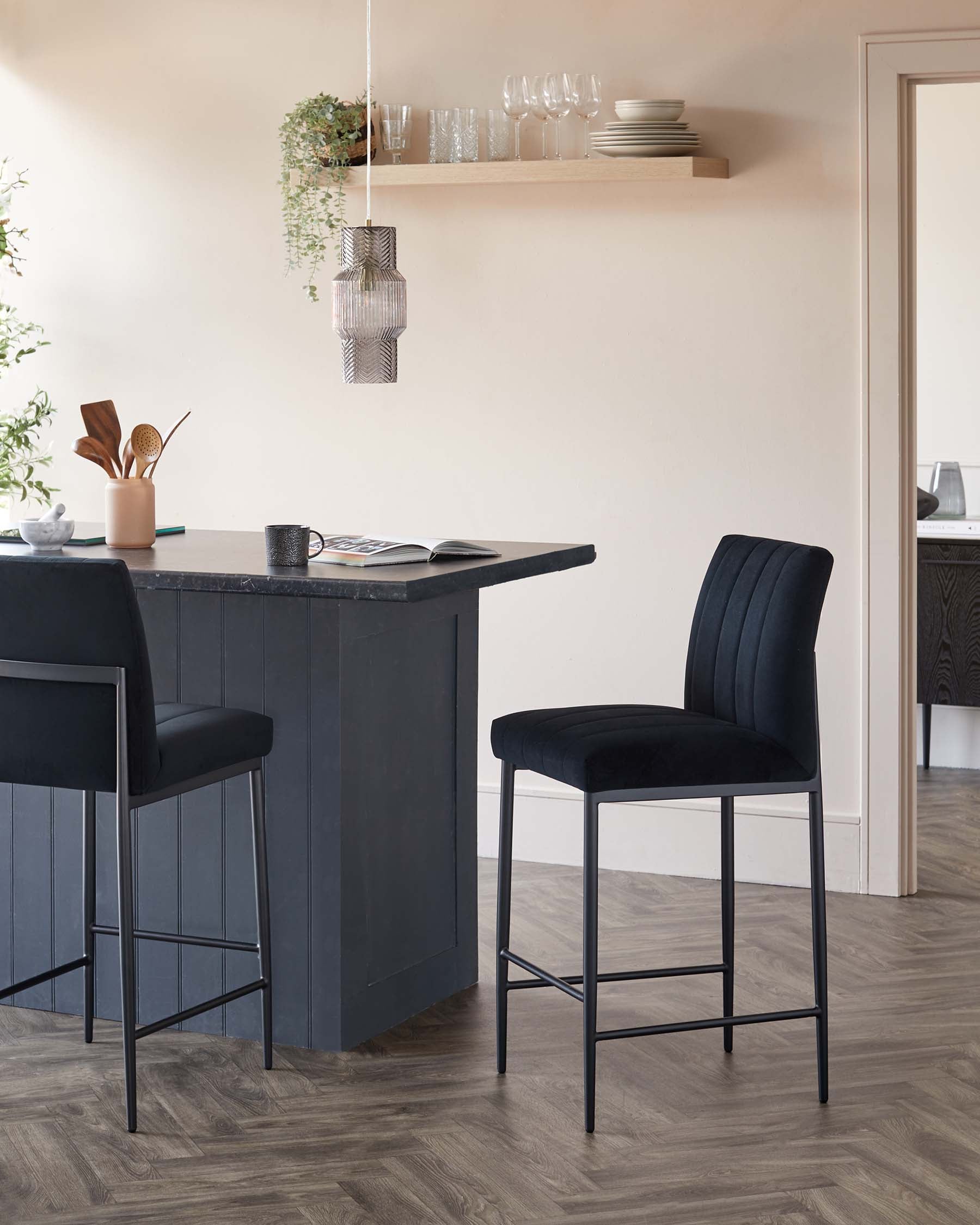 Elegant modern kitchen interior featuring a dark grey kitchen island with a black countertop and panelling details, accompanied by two sophisticated bar stools with black upholstered seats and backs, and sleek metal legs. A floating natural wood shelf displays a simple arrangement of glassware and ceramic dishes above the island.