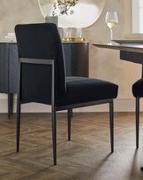 Elegant black upholstered dining chair with slim metal legs and a rectangular dining table with a dark wood finish and angled edges, standing on a herringbone-patterned wooden floor. A decorative sideboard with a textured front and a round mirror above it are partially visible in the background.