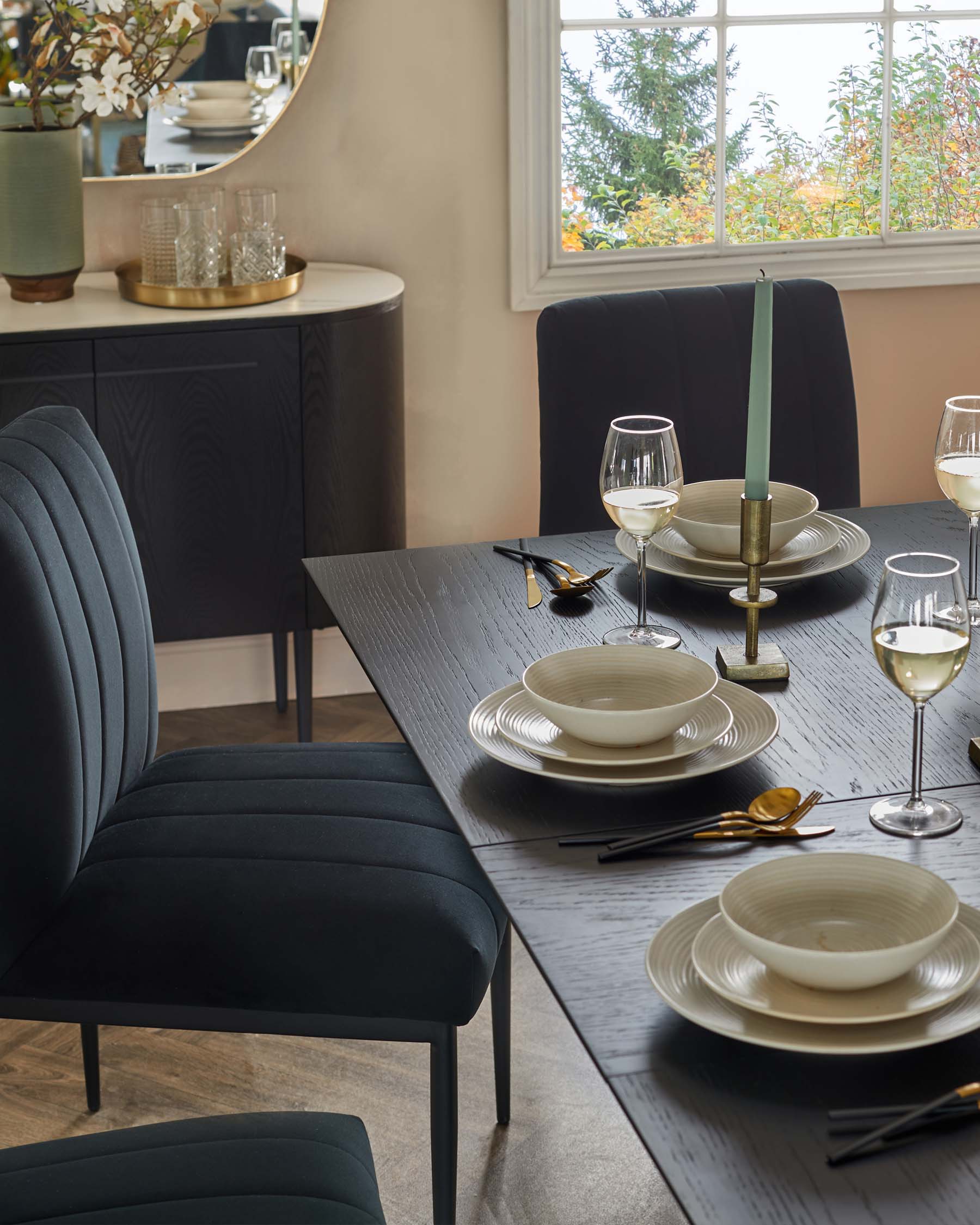 A contemporary dining room featuring an elegant dark wood table, matched with plush, vertical channel-back chairs in dark upholstery. In the background, there's a sleek, curved-edge sideboard with textured front panels, complemented by modern tableware and decorative items.