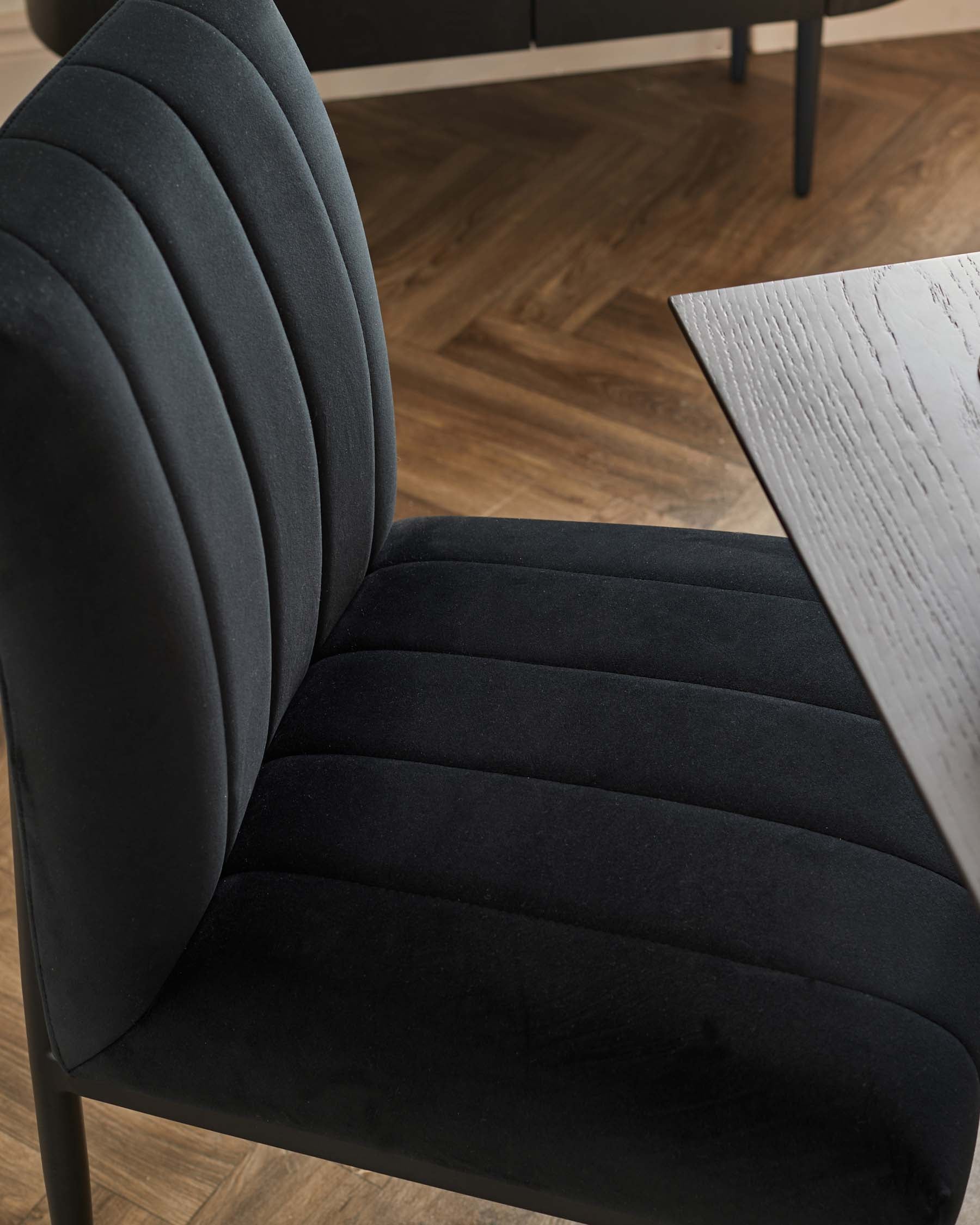 Modern dining chair with vertical channel tufting in black velvet upholstery, featuring a curved backrest and a cushioned seat for comfort, paired with a sleek, black metal frame. A section of a dining table with a textured, white surface and dark legs is also partially visible to the right.