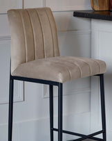 Elegant contemporary bar stool with a high-back design, upholstered in a soft taupe fabric featuring vertical stitching. It stands on a slender, matte black metal frame with four legs and footrests.