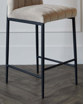 Modern bar stool with a beige upholstered seat featuring channel tufting and a matte black metal frame with footrest, displayed against a white wall on a wood-grain floor.