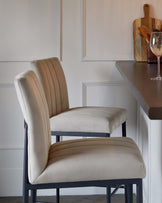 Elegant modern bar stool with a beige upholstered seat and backrest featuring vertical channel tufting and a black metal frame, positioned at a grey countertop.