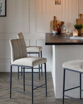 Elegant modern bar stools with beige velvet upholstery and channel tufting on the backrest, seated on a sleek black metal frame with a footrest, positioned adjacent to a kitchen island with a dark countertop.