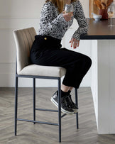 Elegant modern bar stool with a beige upholstered seat and backrest, featuring vertical stitching details. The frame and legs are made of sleek black metal, providing sturdy support. A metal footrest connects the four legs for added comfort. The design is suitable for contemporary kitchen or bar settings.