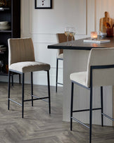 Modern barstools with beige upholstered seats and backs, featuring sleek black metal legs, positioned at a dark stone countertop kitchen island.