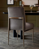 Elegant taupe dining chair with sleek metal legs and a high back, positioned in a sophisticated room setting with wood flooring.