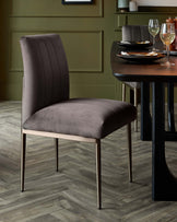 Elegant modern dining chair with vertical stitching on the backrest and a brownish-grey upholstered seat, standing on slim, straight metal legs with a gold finish. Partly visible round dining table with a dark stained wooden top and black legs, complemented by a set for two with white plates, gold cutlery, and stemmed wine glasses. The setting is accented by a green wall with moulding and a wooden floor with a herringbone pattern.