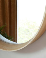 Round wooden mirror with a simple, natural finish frame, mounted on a wall near a window with a glimpse of greenery outside.