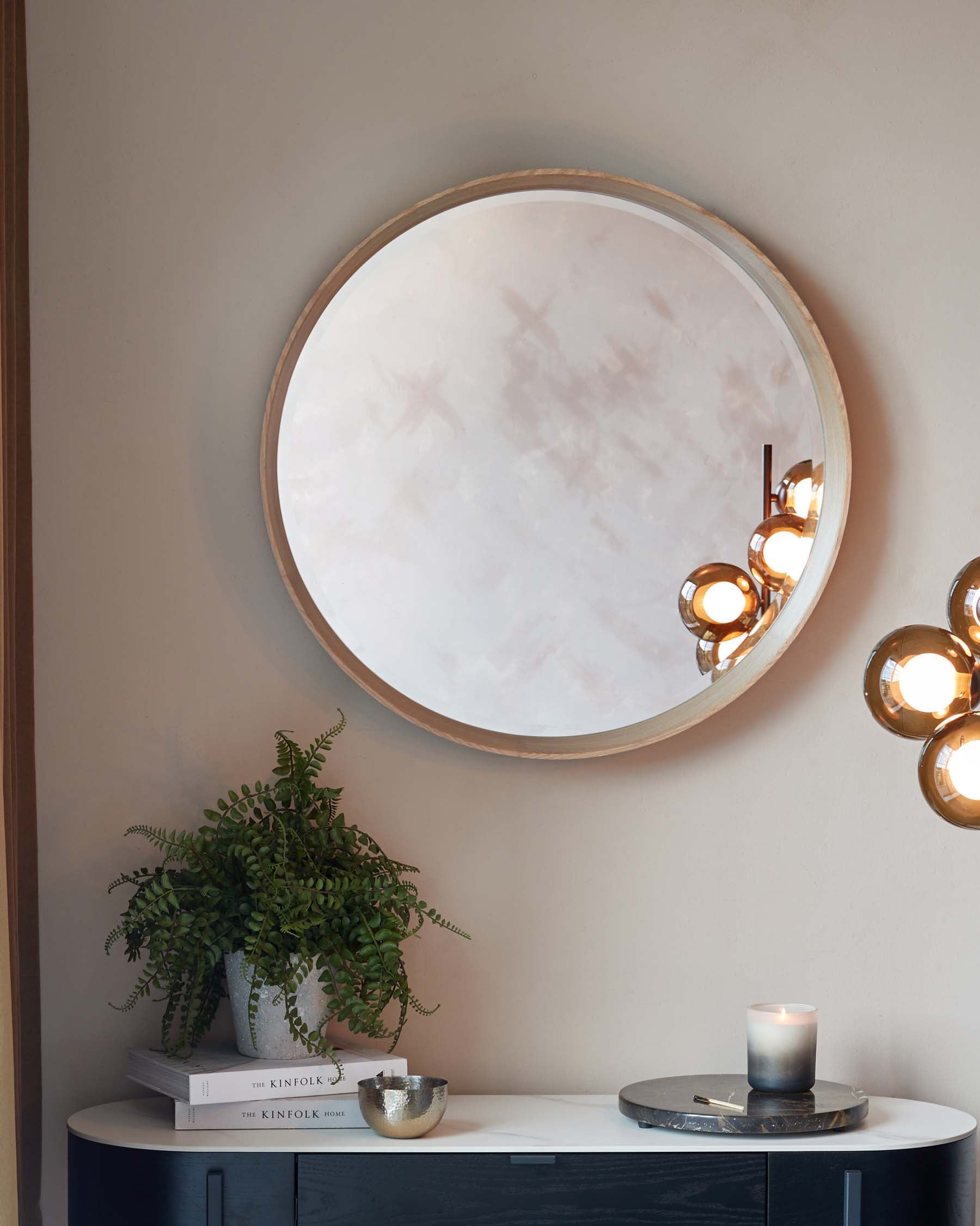 Elegant modern console table featuring a white tabletop with rounded edges and a sleek black base, styled with decorative items such as books, a potted plant, and candles for a minimalist yet warm ambiance. The table is positioned under a large round mirror with a slim wooden frame, enhancing the room's sense of space.