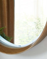 Round wooden mirror with a sleek, natural finish mounted on a white wall near a window with a view of green foliage.