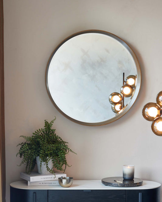 A sleek, modern console table with a semi-circular top in a high-gloss white finish, supported by a dark, charcoal-grey base with cabinet doors. Above the table hangs a large circular mirror with a minimalistic thin, metallic frame. The table is accessorized with a potted fern, a stack of books, and decorative items including a candle and a small bowl.