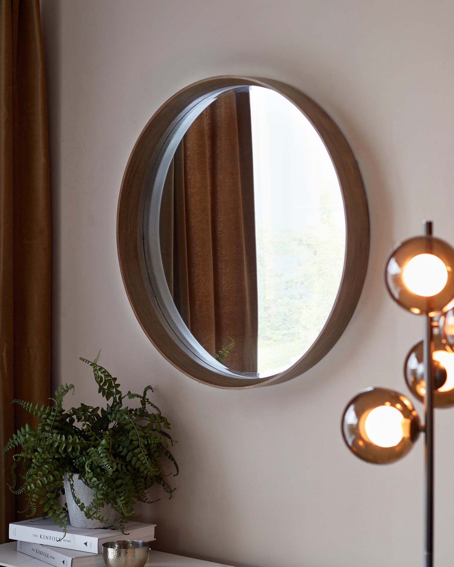 A sleek modern side table with a white top displaying a potted fern, a stack of hardcover books, and a small decorative item. The table is featured next to a wall with a large round mirror framed in a dark wooden finish, complementing the refined contemporary aesthetic of the space.