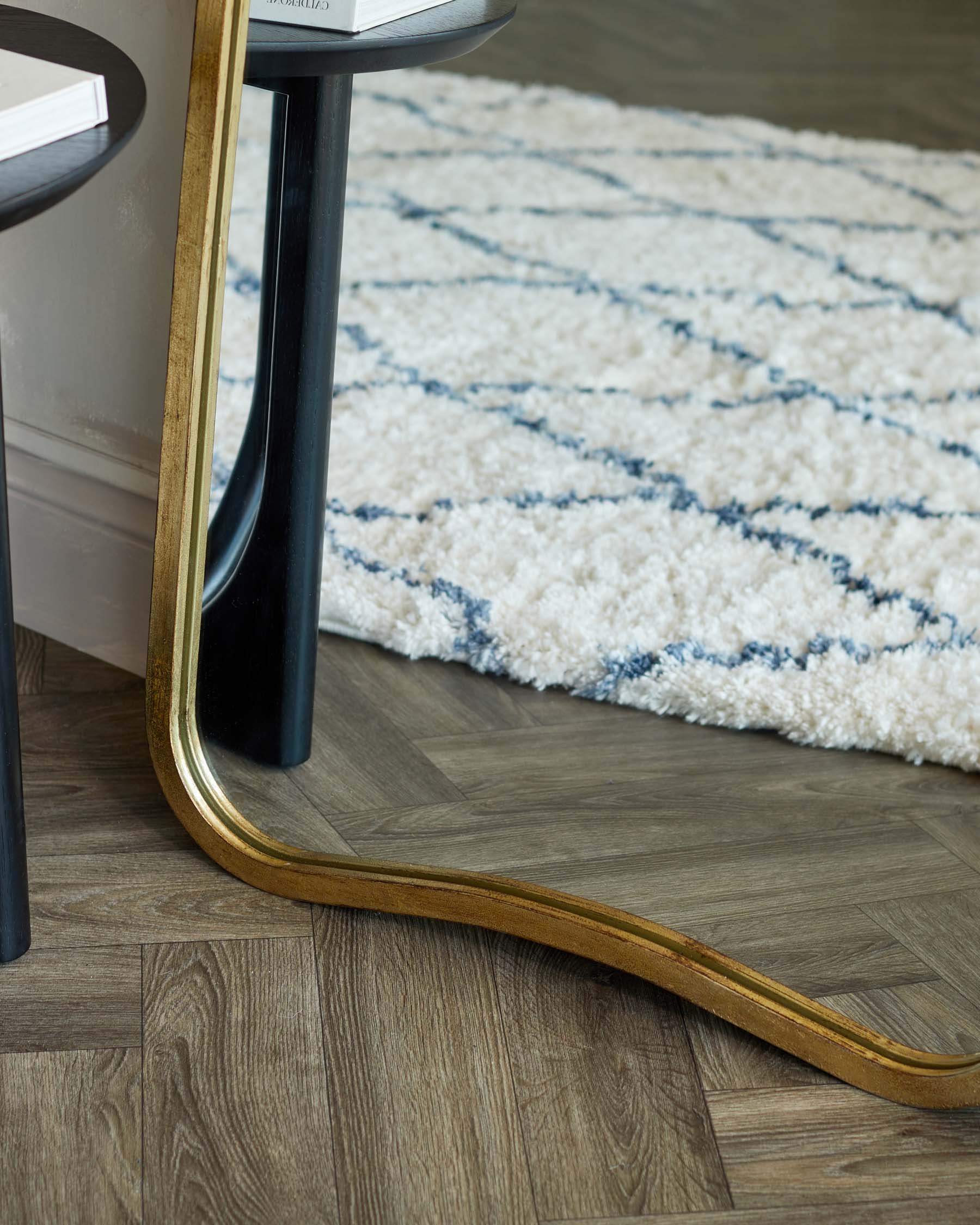 A modern side table with a round marble top supported by a central black pillar with golden accents and a curved, gold-coloured metallic base, resting on a wood-patterned floor beside a plush white and blue patterned area rug.