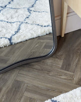 Part of a light wooden bedside table with a leg visible, on hardwood flooring beside a plush white area rug with blue geometric patterns and a grey edge trim.