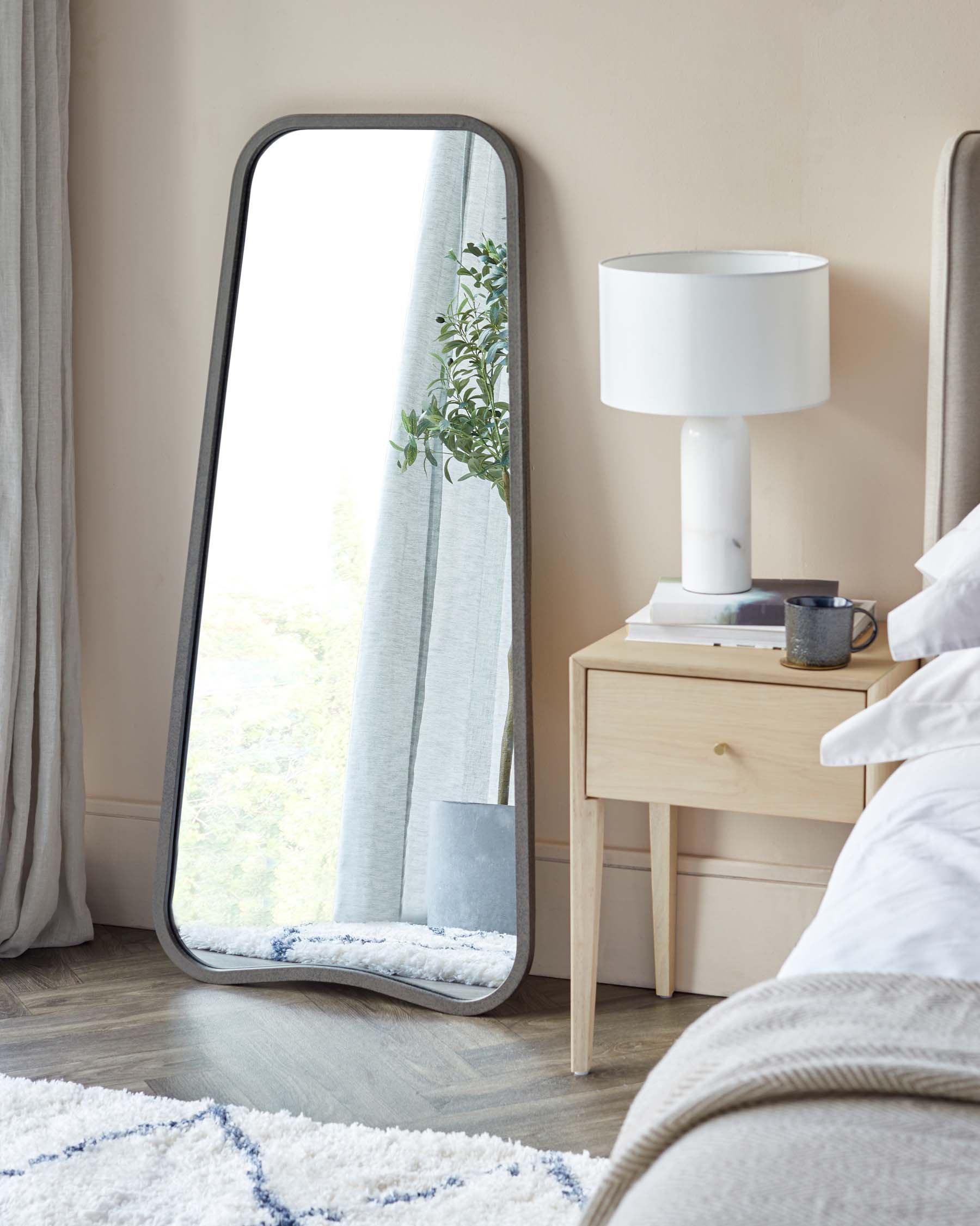 A modern Scandinavian-style bedroom featuring a light wooden bedside table with a drawer and shelf, paired with a sleek white table lamp with a cylindrical shade. A full-length floor mirror with a black frame leans against the wall, reflecting a glimpse of the outdoor view and interior drapery. Elements of cosy decor include a plush white area rug with blue accents on the floor.