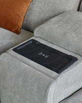 A modern sofa with plush grey upholstery, featuring a sleek wireless charging tray on the armrest and a contrasting orange pillow.