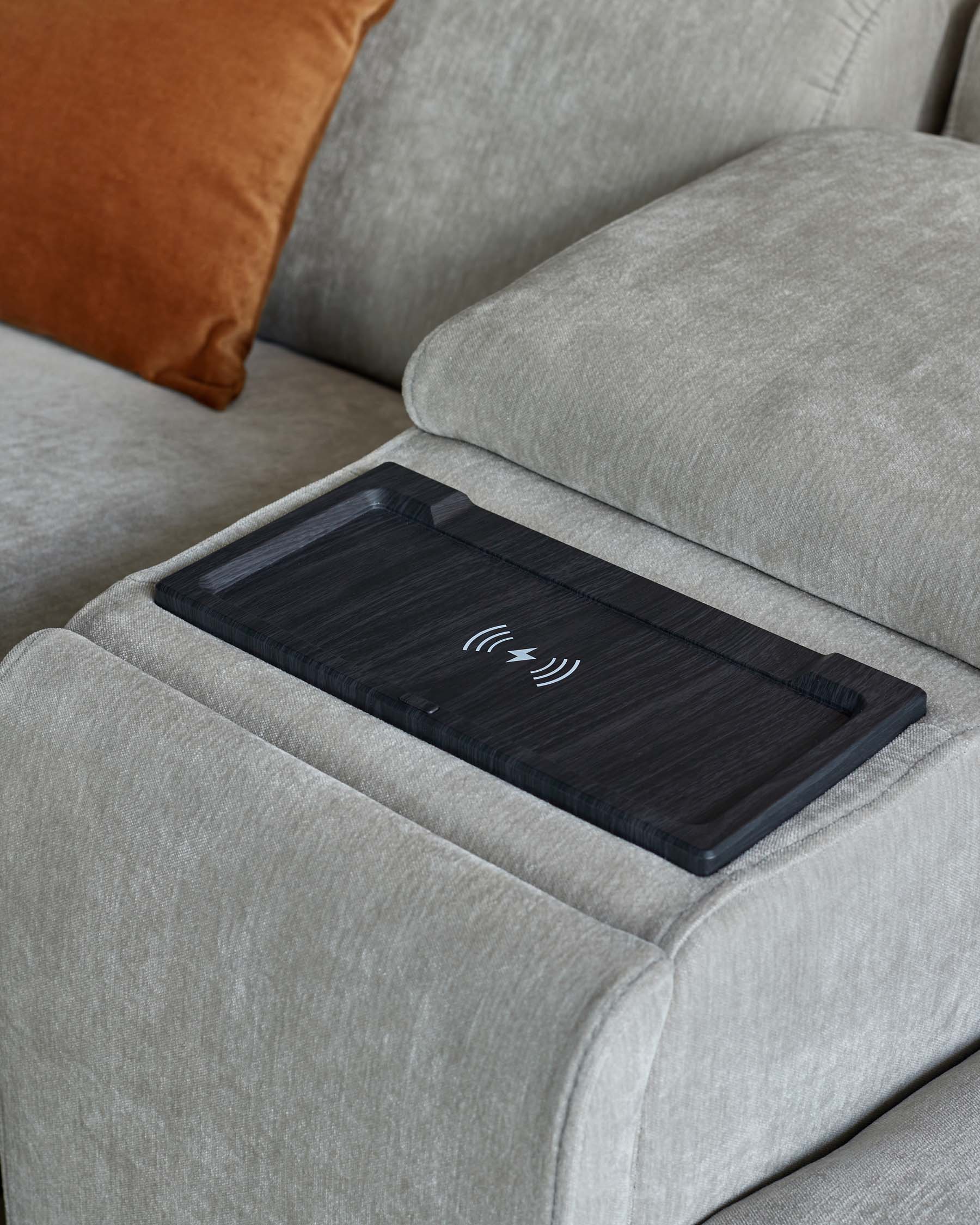 A modern sofa with plush grey upholstery, featuring a sleek wireless charging tray on the armrest and a contrasting orange pillow.