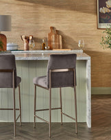 A set of two modern bar stools with grey velvet upholstery and slender, gold-finished metal legs positioned at a marble-topped bar table with a green panelled facade. The stools feature a minimalistic design with a square seat and no backrest, offering a sleek and contemporary look suitable for a stylish kitchen or bar area.