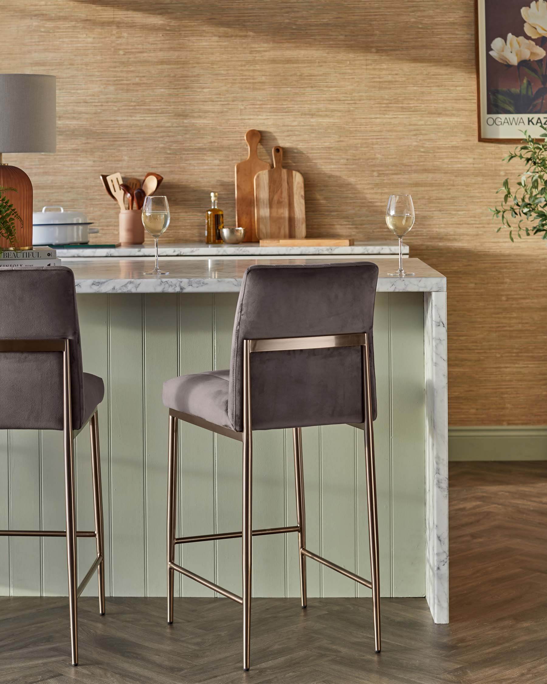 A set of two modern bar stools with grey velvet upholstery and slender, gold-finished metal legs positioned at a marble-topped bar table with a green panelled facade. The stools feature a minimalistic design with a square seat and no backrest, offering a sleek and contemporary look suitable for a stylish kitchen or bar area.